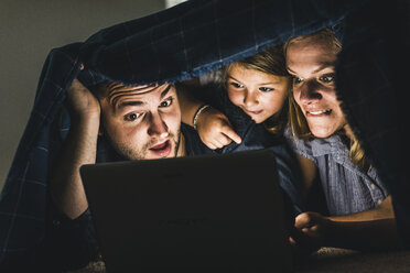 Happy family hiding under blanket, watching film on laptop - UUF11822