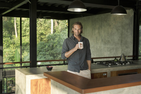 Gut aussehender Mann trinkt Kaffee in einer modernen Design-Küche mit Glasfassade, umgeben von einem üppigen tropischen Garten, lizenzfreies Stockfoto