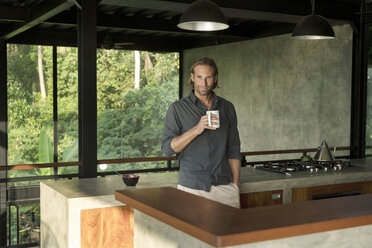 Gut aussehender Mann trinkt Kaffee in einer modernen Design-Küche mit Glasfassade, umgeben von einem üppigen tropischen Garten - SBOF00800