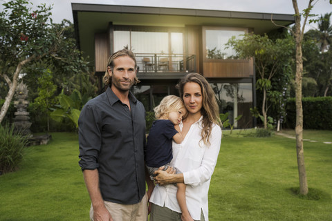 Porträt einer lächelnden Familie, die vor ihrem Designhaus inmitten eines üppigen tropischen Gartens steht, lizenzfreies Stockfoto