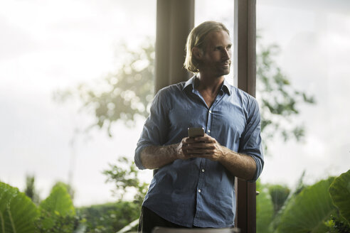 Gut aussehender Mann mit Smartphone in einem modernen Designhaus mit Glasfassade, umgeben von einem üppigen tropischen Garten - SBOF00813