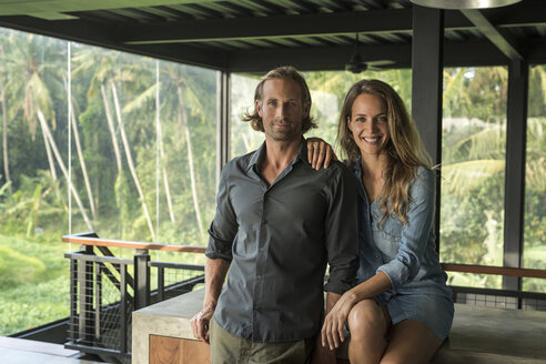 Paar lächelt in die Kamera in einem modernen Designhaus mit Glasfassade, umgeben von einem üppigen tropischen Garten - SBOF00817