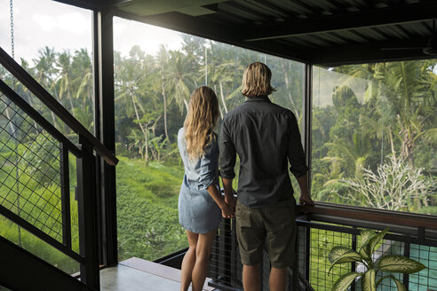 Paar mit Blick auf atemberaubende Aussicht durch Glasfassade in zeitgenössischen Design-Haus von üppigen tropischen Garten umgeben - SBOF00818