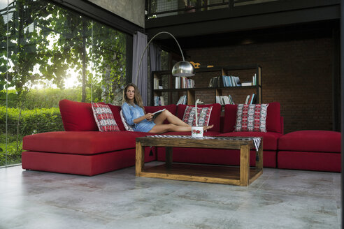 Frau mit Buch entspannt auf roter Couch in modernem Wohnzimmer mit Glasfassade - SBOF00819