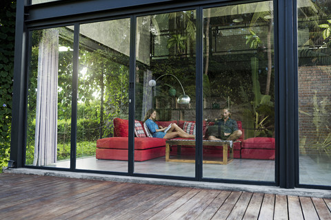 Garden view through glass facade of couple with laptop and book relaxing on red couch in modern design house stock photo