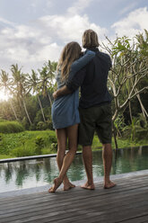 Hugging couple standing on wooden terrace in front of pool and enjoying stunning view of sunset in lush tropical garden - SBOF00826