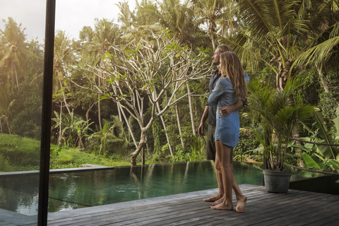 Hugging couple standing on wooden terrace in front of pool and enjoying stunning view in lush tropical garden stock photo