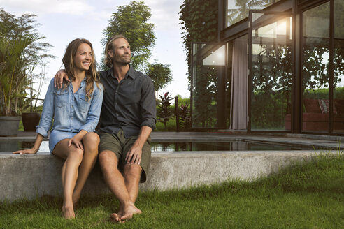 Lächelndes, sich umarmendes Paar, das am Rande eines Pools vor seinem Designhaus sitzt - SBOF00828
