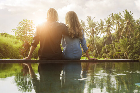 Umarmung Paar sitzt auf dem Rand eines Pools und genießen den atemberaubenden Blick auf den Sonnenuntergang in üppigen tropischen Garten - SBOF00829