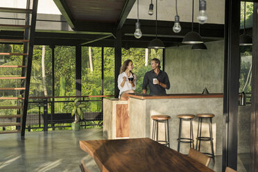 Couple having breakfast and smiling at each other in modern design kitchen with glass facade surrounded by lush tropical garden - SBOF00831