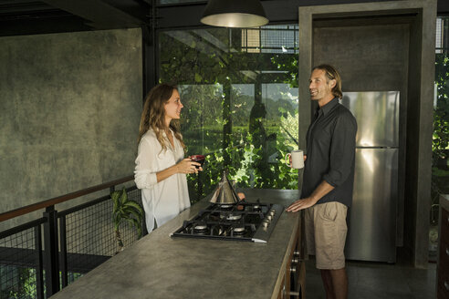 Ein Paar frühstückt und lächelt in einer modernen Designküche mit Glasfassade, umgeben von einem üppigen tropischen Garten - SBOF00833