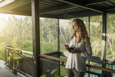 Smiling woman eating cereal in contemporary design house with pool and glass facade surrounded by lush tropical garden - SBOF00834