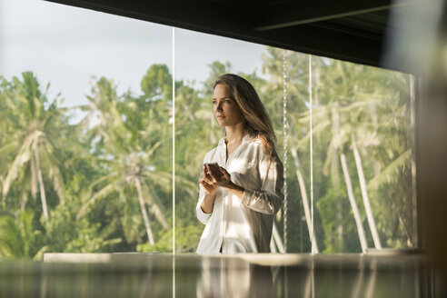 Frau hält Smartphone in einem Designhaus vor einem üppigen tropischen Garten - SBOF00836