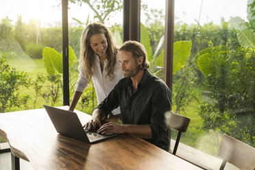 Woman smiling at husband working on laptop in design house surrounded by lush tropical garden - SBOF00839