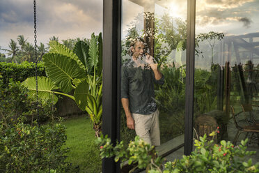 gut aussehender Mann steht hinter der Glasfassade eines Designhauses und spricht mit seinem Smartphone, umgeben von einem üppigen tropischen Garten - SBOF00844