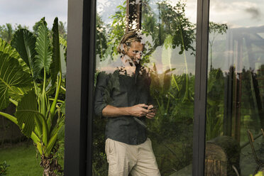 Handsome man standing behind glass facade of design house looking at smartphone surrounded by lush tropical garden - SBOF00845