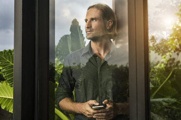 Gut aussehender Mann, der hinter der Glasfassade eines Designhauses steht und ein Smartphone hält, umgeben von einem üppigen tropischen Garten - SBOF00846