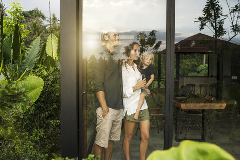 Gartenansicht von Eltern mit ihrem kleinen Sohn, die nach draußen auf ihr Designhaus schauen, das von einem üppigen tropischen Garten umgeben ist - SBOF00847