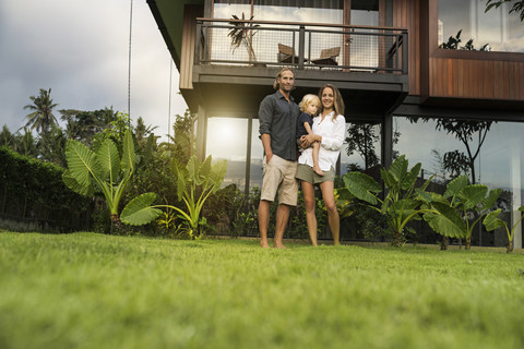 Porträt einer lächelnden Familie, die vor ihrem Designhaus inmitten eines üppigen tropischen Gartens steht, lizenzfreies Stockfoto