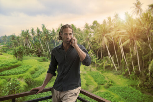 Gut aussehender Mann, der sich auf einen Balkon stützt und mit seinem Smartphone spricht, mit Blick auf eine tropische Landschaft - SBOF00850