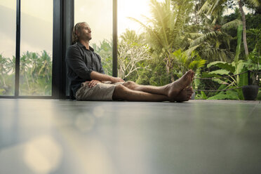 Handsome man sitting on floor and leaning on glass facade with stunning tropical garden in background - SBOF00852