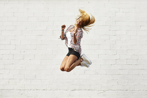 Happy young woman jumping mid-air in front of white wall - JRFF01448