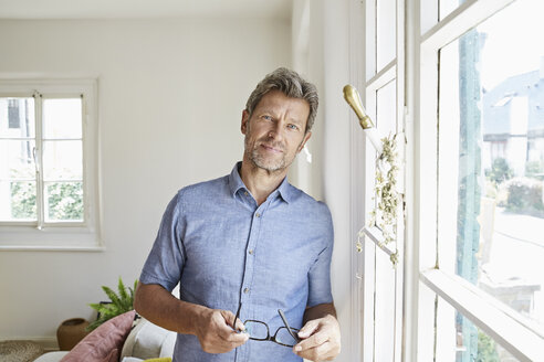 Mature man at home eaning at window, holding glasses - PDF01296