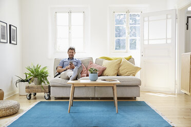 Man sitting on couch at home, using smartphone - PDF01297