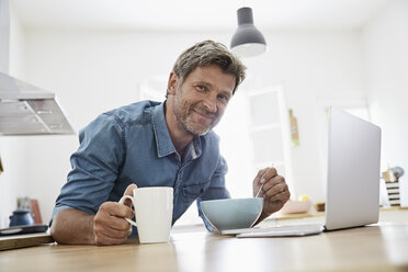 Älterer Mann in seiner Küche beim Frühstück, während er seinen Laptop überprüft - PDF01362