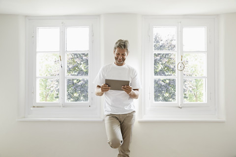 Älterer Mann steht am Fenster und benutzt ein digitales Tablet, lizenzfreies Stockfoto