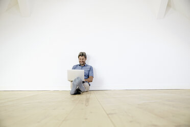 Mature man sitting on ground in empty room, using laptop - PDF01385