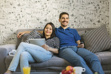 Couple sitting on the couch using tablet and watching television - MOMF00250