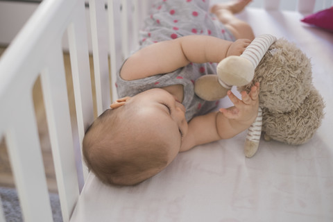 Niedliches Baby-Mädchen in der Krippe liegend und mit Kuscheltier spielend, lizenzfreies Stockfoto