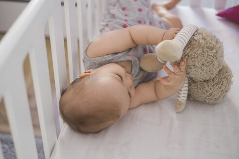 Niedliches Baby-Mädchen in der Krippe liegend und mit Kuscheltier spielend - MOMF00264