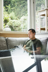 Man at home using tablet on the couch near the window - MOEF00166