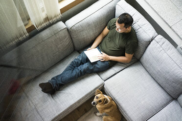 Mann zu Hause mit Tablet auf der Couch mit Hund neben ihm - MOEF00167