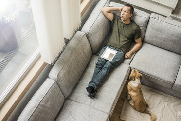 Man at home with tablet on the couch with dog beside him - MOEF00168