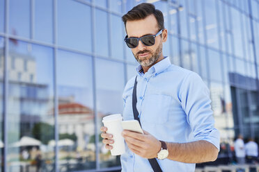 Businessman using phone and holding coffee outdoors - BSZF00029
