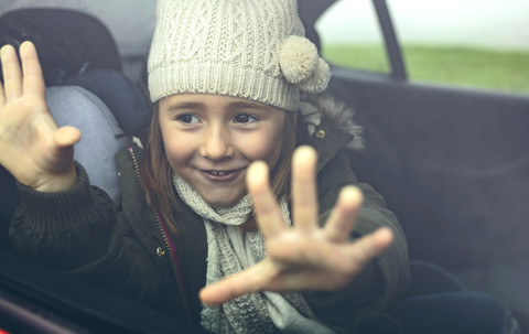 Porträt eines glücklichen kleinen Mädchens mit Wollmütze, das seine Finger auf das Autofenster legt, lizenzfreies Stockfoto