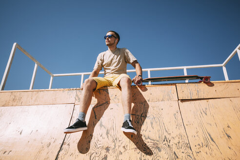 Junger Mann mit Ohrstöpseln und Longboard sitzt auf der Halfpipe im Skatepark - VPIF00213