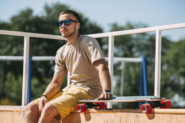 Junger Mann mit Ohrstöpseln und Longboard im Skatepark sitzend - VPIF00214