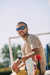 Smiling young man with earbuds and longboard in skatepark - VPIF00215