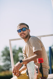 Lächelnder junger Mann mit Ohrstöpseln und Longboard im Skatepark - VPIF00215