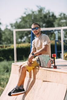 Lächelnder junger Mann mit Ohrstöpseln und Longboard auf der Halfpipe im Skatepark sitzend - VPIF00216