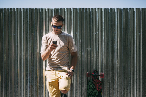 Junger Mann mit Handy und Ohrstöpseln lehnt an einer Wand neben einem Longboard - VPIF00221