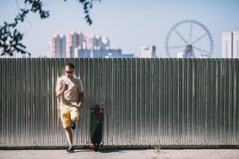 Junger Mann mit Handy lehnt an einer Wand neben Longboard mit Stadt im Hintergrund, lizenzfreies Stockfoto