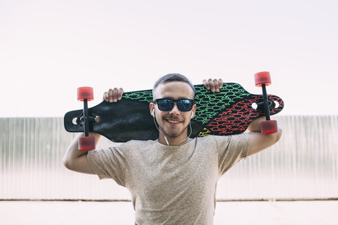 Portrait of confident young man with earbuds carrying longboard - VPIF00230