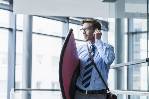 Businessman with earphones carrying surfboard in office stock photo