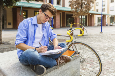 Junger Mann mit Rennrad sitzt auf einer Bank und schreibt auf einem Notizblock - MGIF00140