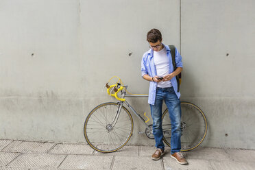 Junger Mann mit Rennrad, der vor einer Betonwand auf sein Handy schaut - MGIF00150